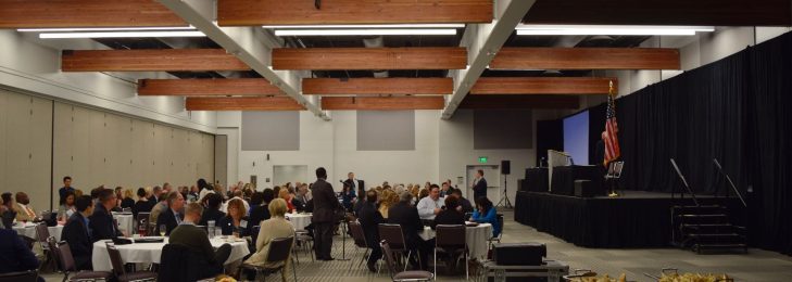 Inside of a building during a conference, with visitors gathered at tables looking at a stage.
